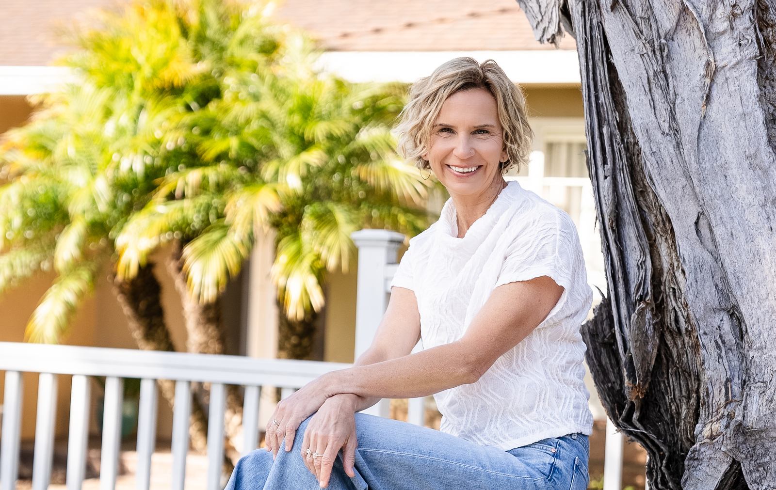 Michelle sitting against a palm trunk in the sun, smiling, garden and white fence behind her